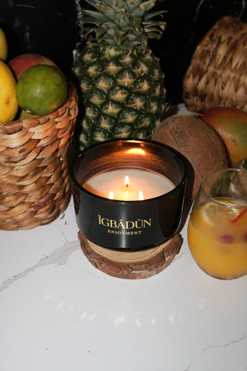 A black bowl candle with a gold label, featuring multiple organic wicks, burning with a warm glow. The candle is made with natural coconut and soy wax and non toxic fragrance oils